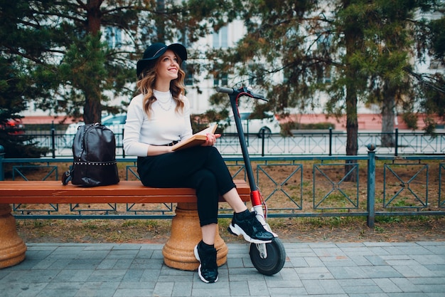 Anuncio de libro de lectura de mujer joven sentada en un banco con scooter eléctrico en la ciudad