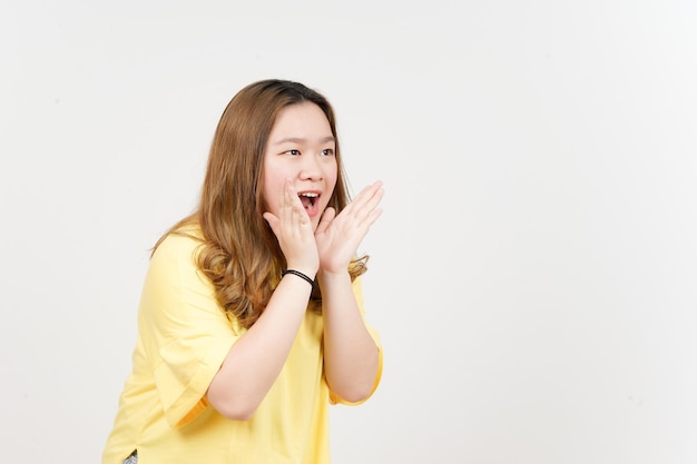 Anuncio con las manos sobre la boca de una hermosa mujer asiática con camiseta amarilla aislada en blanco