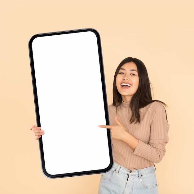 Anúncio móvel sorrindo mulher asiática segurando e apontando para um grande smartphone em branco