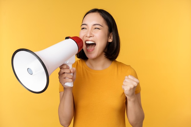 Anuncio Mujer asiática feliz gritando fuerte en megáfono reclutando protestando con altavoz en manos de pie sobre fondo amarillo