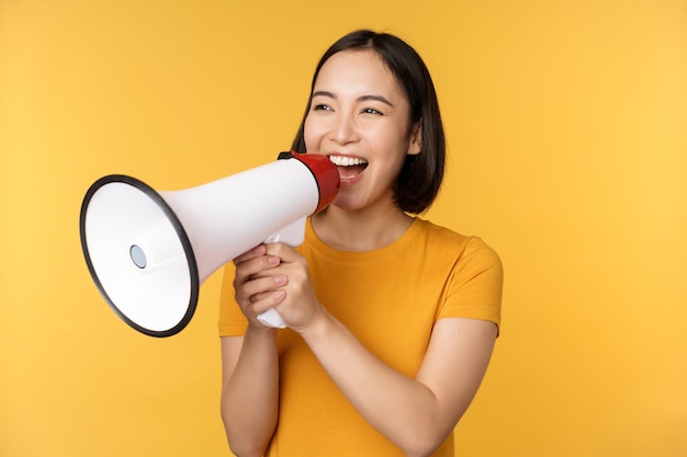 Anúncio mulher asiática feliz gritando alto no recrutamento de megafone protestando com alto-falante nas mãos sobre fundo amarelo