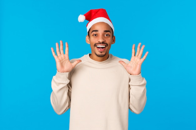 Anziehende Geschenke des Mannes in Sankt-Hut. Der nette junge Kerl des Afroamerikaners ergreifen etwas, heben Hände an und warten auf den Fang und froh lächeln, feiern Winterurlaube, blaue Wand