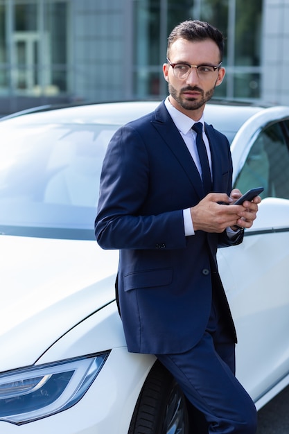 Anzug und Krawatte. Geschäftsmann in Anzug und Krawatte mit Smartphone, während er in der Nähe des Autos steht