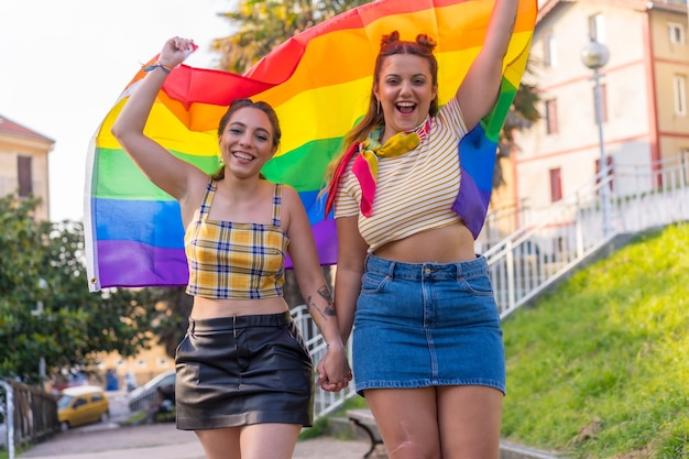 Ao ar livre, uma foto de duas jovens caucasianas se abraçando com a bandeira do orgulho LGBT