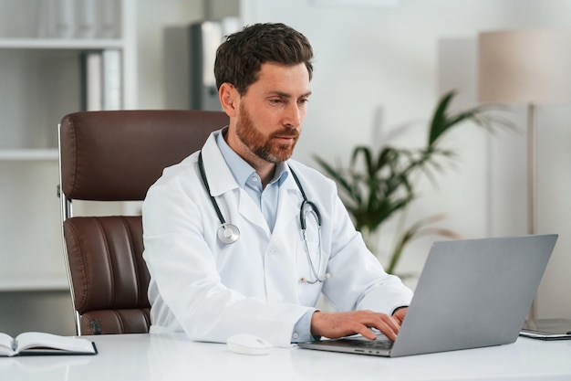 Foto ao lado da mesa com o portátil, o médico de roupa formal está a trabalhar no gabinete.