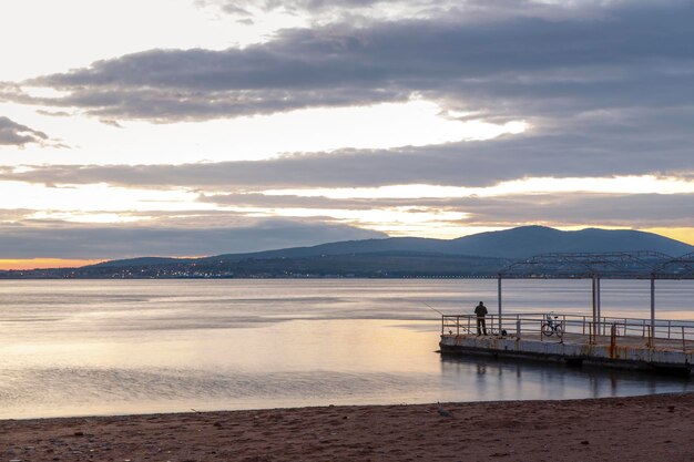 Ao pôr do sol, um pescador está parado com uma vara de pescar em um píer de concreto. Nas montanhas ao fundo