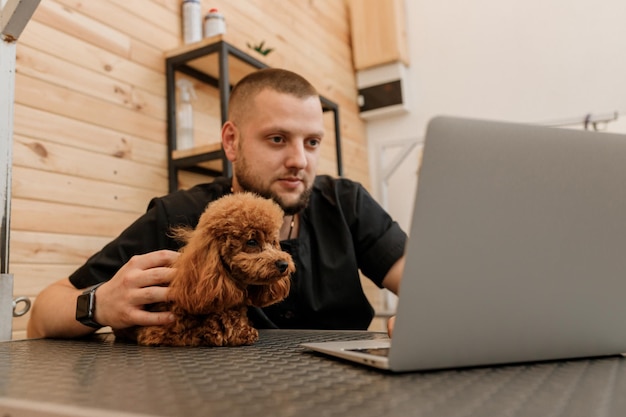 Aparador masculino profissional trabalhando no laptop em seu local de trabalho no salão de beleza perto com o cão poodle Conceito de aliciamento de animais