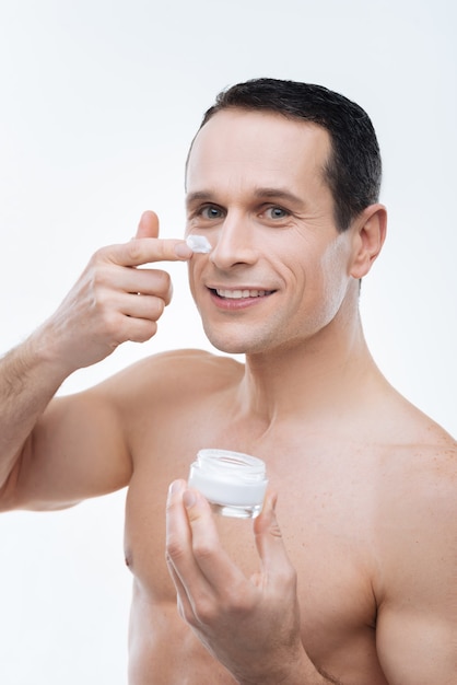 Aparência perfeita. Homem alegre e positivo sorrindo e usando creme facial enquanto se preocupa com sua aparência