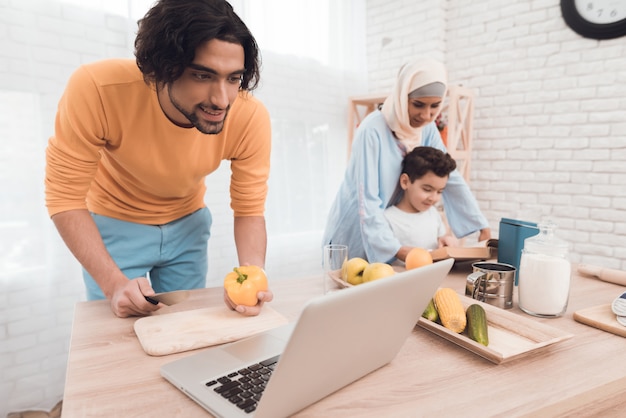 Apariencia árabe en ropa moderna en cocina con laptop.