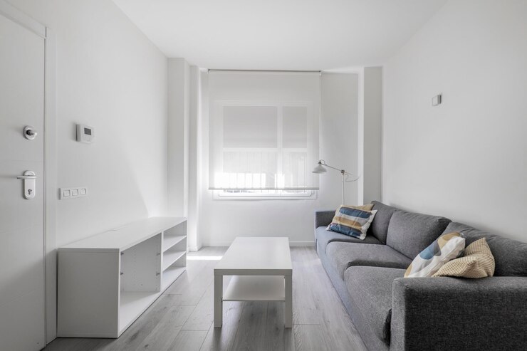 Foto apartamento com sala em tons de branco e sofá cinza com piso de madeira clara e janela com persiana