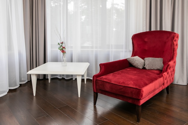 Apartamento de luxo com sofá vermelho de estilo clássico, piso de madeira, mesa de café decorada com vaso de flores e grande janela panorâmica com cortinas transparentes