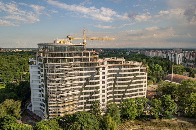 Apartamento ou prédio alto em construção. Paredes de tijolo, janelas de vidro, andaimes e pilares de suporte de concreto. Guindaste de torre no espaço da cópia do céu azul brilhante