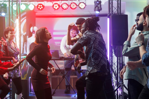 Apasionada pareja multirracial bailando juntos y de fiesta en la discoteca. Despreocupados hombres y mujeres clubbers haciendo movimientos al ritmo de la música en la pista de baile iluminada con focos de escenario