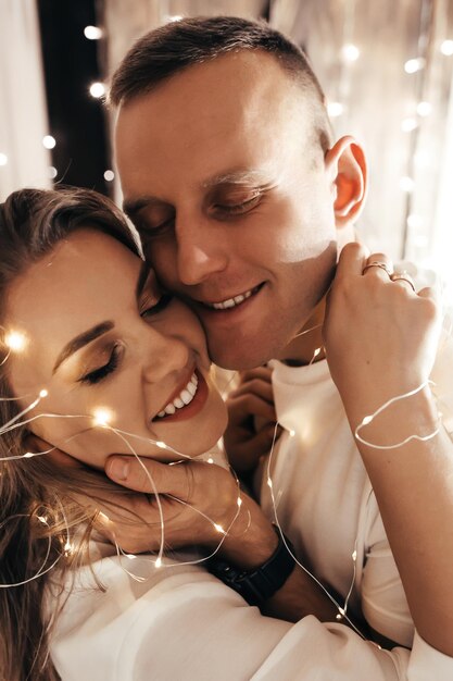 Apasionada pareja romántica pasando tiempo antes de Año Nuevo en casa con guirnaldas. Mujer joven y hombre guapo abrazo, beso. Tiempo de Navidad.