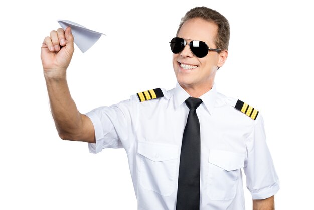 Apasionado de su trabajo. Confiado piloto masculino en uniforme jugando con avión de papel mientras está de pie contra el fondo blanco.