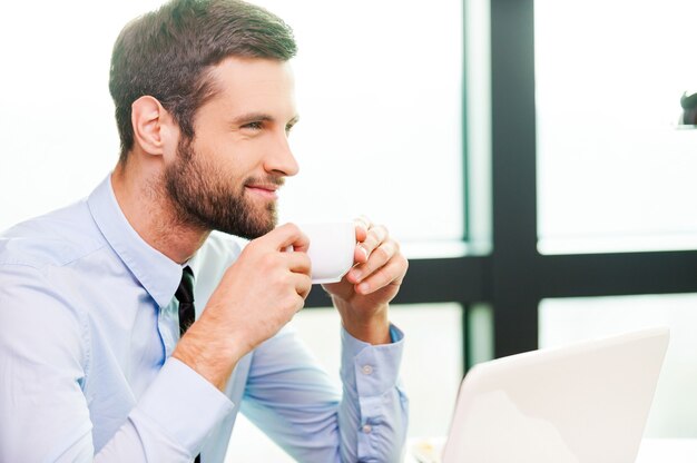 Apenas inspirado. Vista lateral de um jovem pensativo de camisa e gravata, segurando a xícara de café e olhando para longe enquanto está sentado em seu local de trabalho