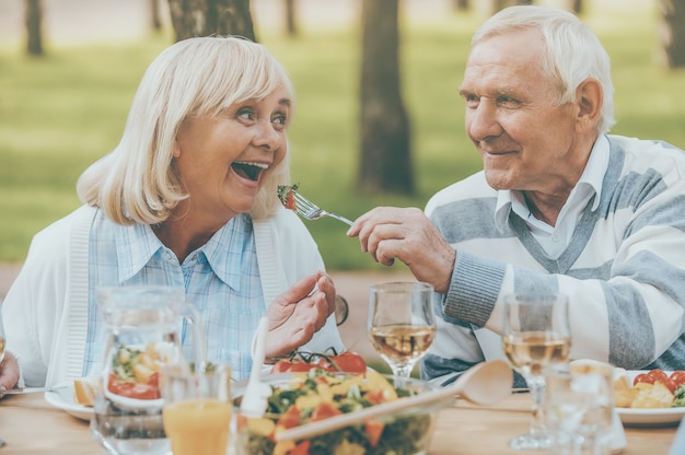 Apenas o melhor para o nosso mais próximo. Último homem alimentando sua alegre esposa com salada fresca, enquanto os dois estão sentados à mesa de jantar ao ar livre