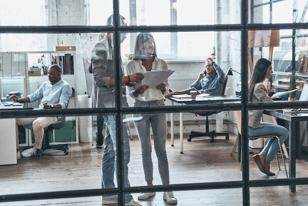 Apenas um dia de trabalho. Comprimento total de jovens modernos trabalhando e se comunicando enquanto estão atrás de uma parede de vidro na sala da diretoria