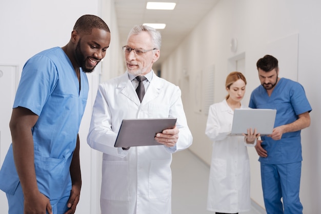 Apenas veja a diferença. pediatra de envelhecimento habilidoso e alegre aproveitando o trabalho no hospital e trabalhando com um tablet, enquanto outros colegas usando o laptop