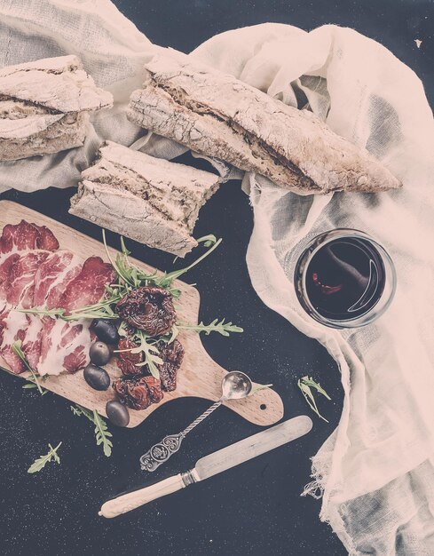 Aperitivo de vinho conjunto de louça vintage baguete francesa quebrada em pedaços tomates secos azeitonas defumadas carne e rúcula na placa de madeira rústica sobre fundo escuro Tom pastel