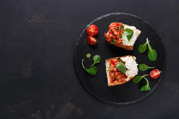 Aperitivo italiano sobre fondo negro. Bruschetta crujiente con tomates concasse, queso stracciatella decorado con espinacas. Comidas de restaurante deliciosas y saludables, vista superior, espacio de copia