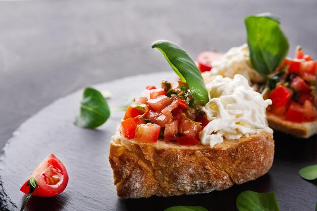 Aperitivo italiano sobre fondo de pizarra negra. Bruschetta crujiente con tomates concasse, queso stracciatella decorado con espinacas. Comidas de restaurante deliciosas y saludables, espacio de copia, primer plano