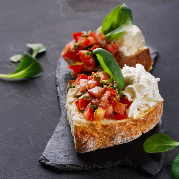 Foto aperitivo de restaurante sobre fondo negro. bruschetta crujiente con tomates concasse, queso stracciatella decorado con espinacas. comidas deliciosas y saludables, copie el espacio, primer plano