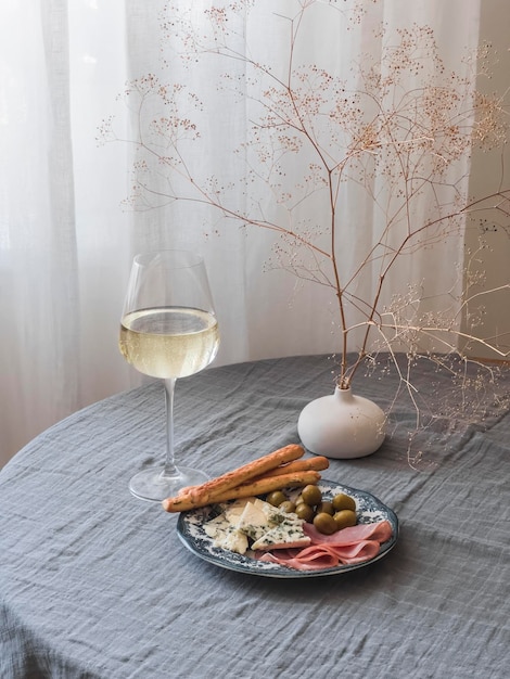 Aperitivo um copo de vinho branco e um prato com queijo, presunto, azeitonas e palitos de pão na mesa