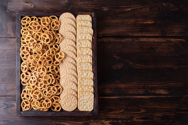 Aperitivos de fiesta, caja con galletas, galletas saladas y mini pretzels salados sobre un fondo de madera oscura, primer plano.