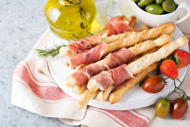 Aperitivos italianos tradicionales para vino, palitos de pan (grissini), tomates, prosciutto (jamón), fresas y aceitunas. Enfoque selectivo.