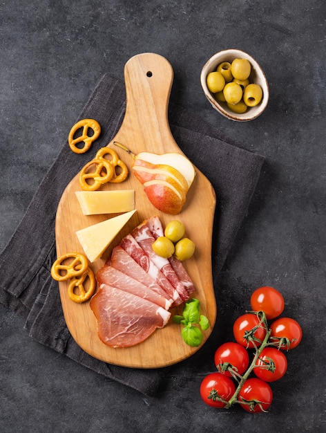 Aperitivos variados o antipasti Tabla de aperitivos Queso parmesano con jamón, aceitunas, pera y galletas saladas sobre fondo oscuro con tomates Vista superior