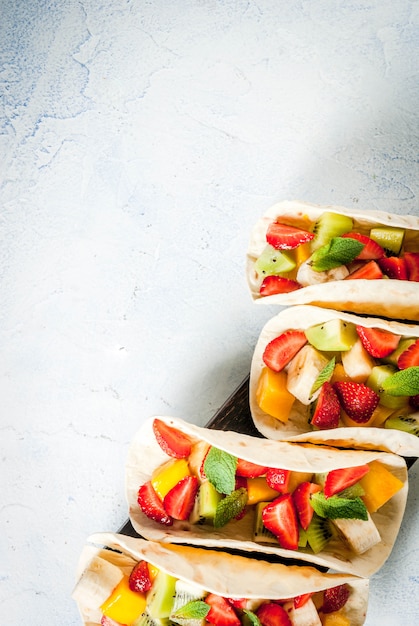 Aperitivos de verano. Comida para una fiesta. Tacos de frutas con fresas, mangos, plátanos, chocolate, menta. Sobre una mesa de hormigón azul claro. vista superior de copyspace