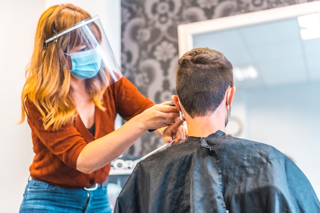 Foto apertura de salones de peluquería después de la pandemia de coronavirus, covid-19. medidas de seguridad, mascarilla, pantalla protectora, distancia social. peluquera trabajando con una joven morena