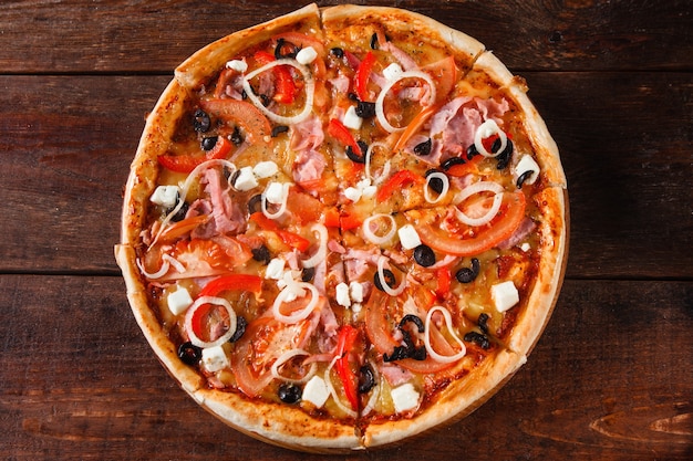 Apetitosa y deliciosa pizza italiana colorida con jamón, tomates, aceitunas, cebolla y queso feta servido en mesa de madera, plano.