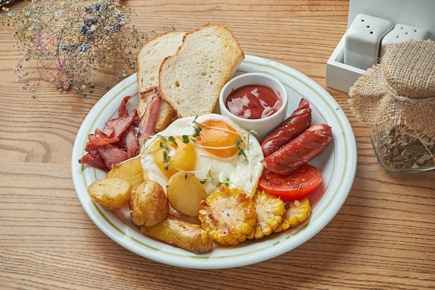 Una apetitosa opción de desayuno: huevo frito con salchichas, tocino y salchichas, papas y maíz, servido en un plato blanco con salsa roja. Comida de restaurante