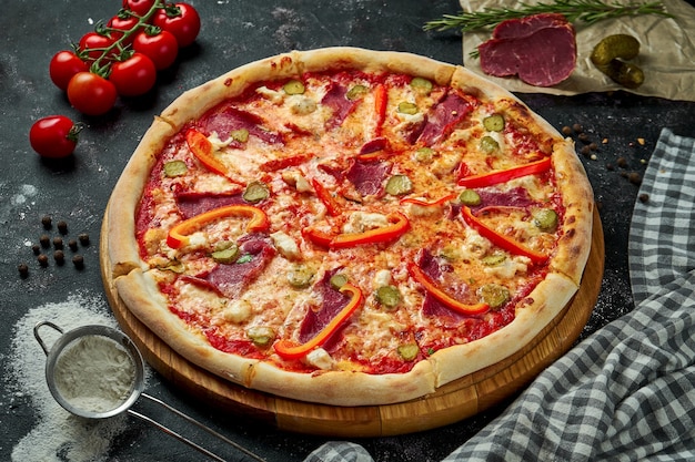 Apetitosa pizza italiana con queso, pimiento, jamón, pepinos encurtidos y salsa roja en una composición con ingredientes sobre una mesa negra