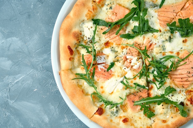 Foto apetitosa pizza de queso con salmón y rúcula en un plato blanco sobre hormigón vista superior.