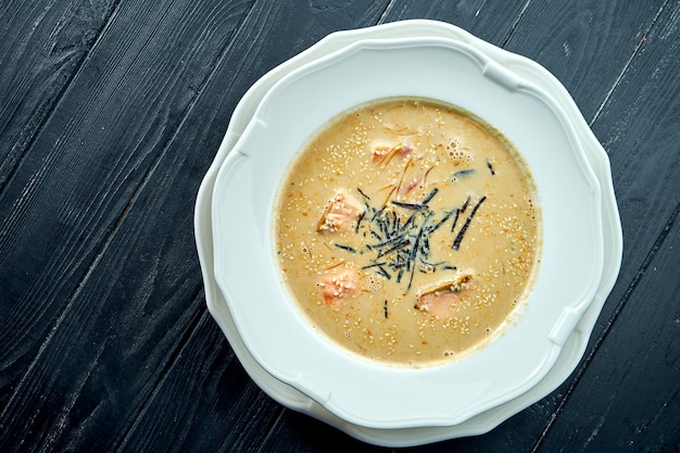 Apetitosa sopa de coco con salmón, nori y semillas de sésamo, servida en un plato blanco sobre una mesa de madera oscura.