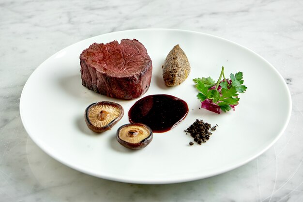 Apetitoso bife mal passado com cogumelos e molho de frutas vermelhas, servido em prato branco sobre mesa de mármore. Comida de restaurante