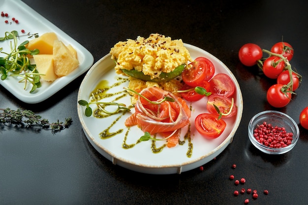 Apetitoso desayuno - huevos revueltos con salmón y aguacate en un plato blanco sobre un fondo oscuro con ingredientes