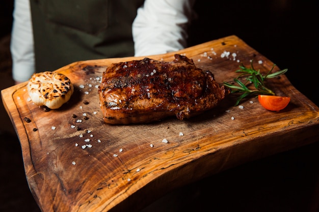 Un apetitoso filete de carne a la parrilla recién preparado se encuentra sobre una tabla de madera. Rociado con trozos de sal, al lado hay una ramita de razmarin y una rodaja de tomate.