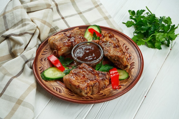 Apetitoso frito costelas de porco assado com molho vermelho e legumes em um prato de cerâmico em um fundo branco de madeira.