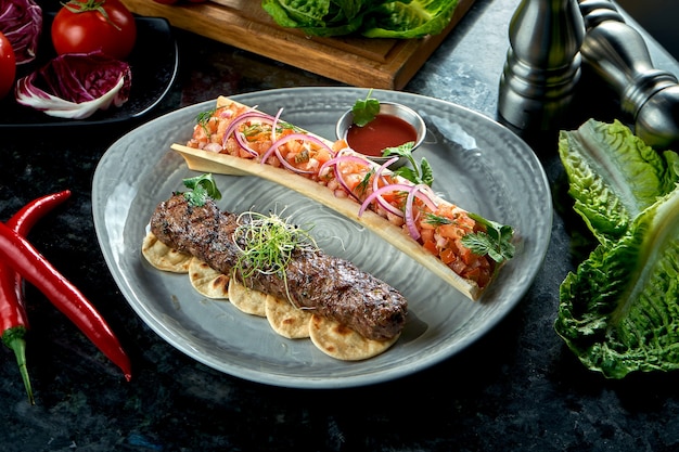 Apetitoso kebab de lula de cordero turco con tartar de tomate y salsa roja, servido en un plato gris. Mesa de mármol oscuro. Carne a la barbacoa, comida de restaurante.