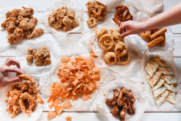 Apetitoso surtido de bocadillos de cerveza fresca en la mesa de madera blanca, endecha plana. Comida chatarra, alimentación poco saludable, calorías. Persona irreconocible tomar piezas, comedor, buffet