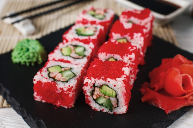 Apetitosos rollos de sushi fresco con cangrejo, pepino y caviar tobiko rojo en un restaurante japonés servido en pizarra negra con jengibre y wasabi, primer plano. Mariscos deliciosos.