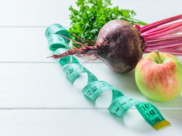 Apfel, Rote Beete, Petersilienstrahl und Maßband auf weißem Holztisch.