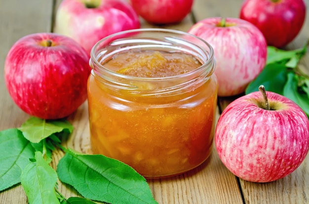 Apfelmarmelade im Glas, frische rote Äpfel, Zweige mit Blättern auf einem Holzbrett