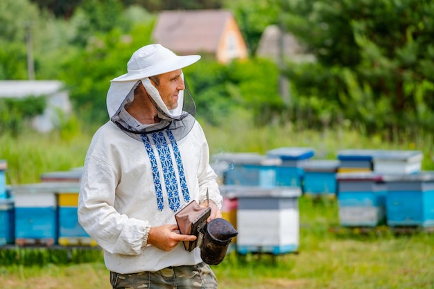 Apicultor en soportes de sombrero protector con fumador para abejas en las manos Colmenas en el fondo Día soleado de verano en el colmenar