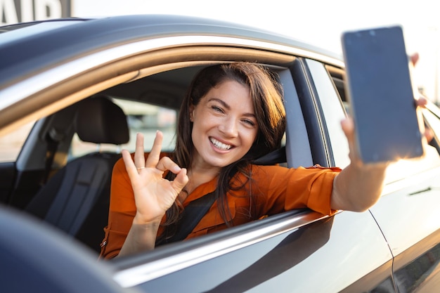 Foto aplicación de alquiler de automóviles mujer feliz mostrando un teléfono inteligente en blanco mientras está sentada dentro de un automóvil mujer sonriente demostrando espacio de copia para publicidad móvil o maqueta de sitio web