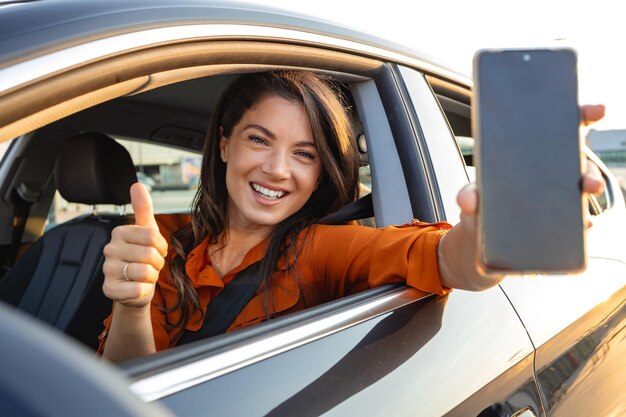 Foto aplicación de alquiler de automóviles mujer feliz mostrando un teléfono inteligente en blanco mientras está sentada dentro de un automóvil mujer sonriente demostrando espacio de copia para publicidad móvil o maqueta de sitio web